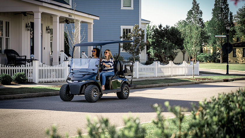 Two women drive an E-Z-GO vehicle down a street.